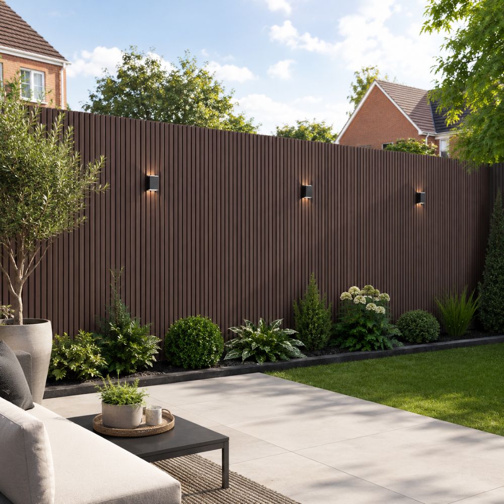 A modern outdoor patio area with a tall dark brown vertical slat privacy fence, green lawn, potted plants, and a light gray stone patio. A partial white sofa and black coffee table are visible in the foreground.