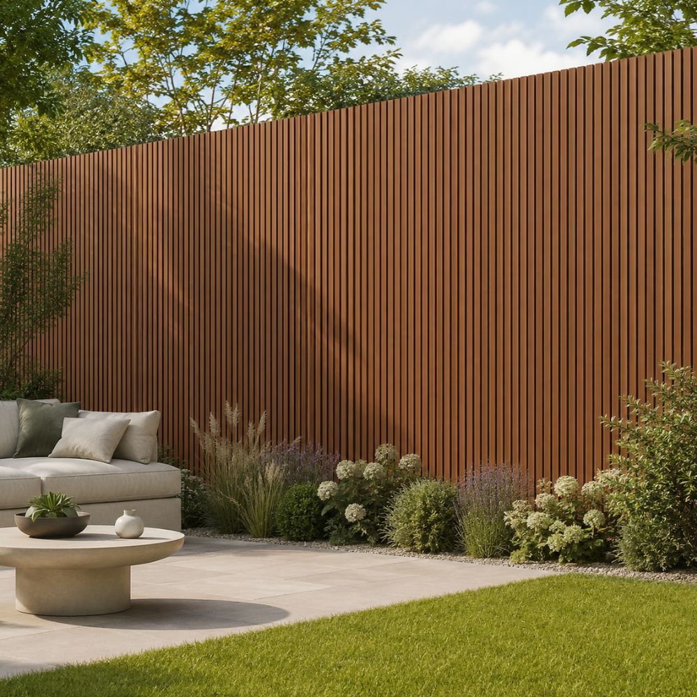 A modern outdoor patio scene with a large vertical slat fence in a warm brown/rust color, landscaped with green shrubs, grass, and a light gray lounge chair with pillows, plus a round stone coffee table in the foreground.