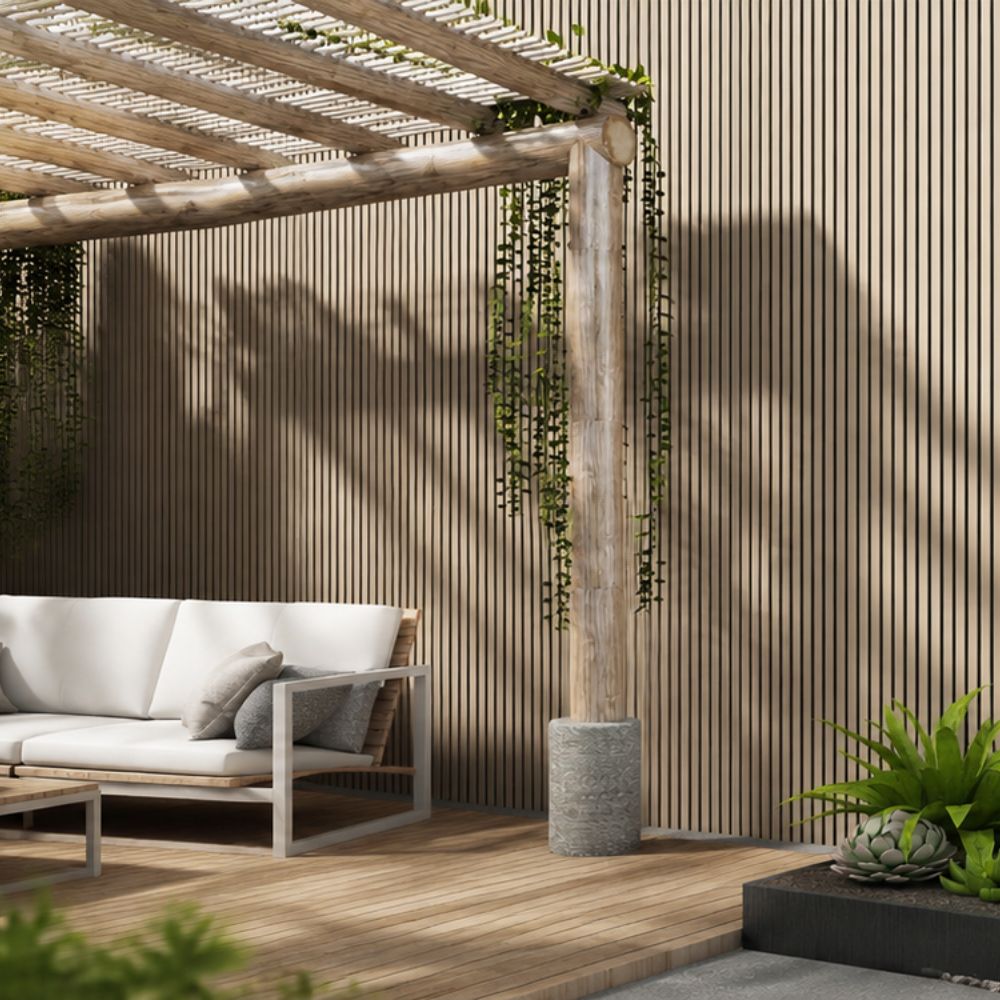 A modern outdoor patio scene with a slatted wood wall, wooden pergola beams, hanging greenery, a white cushioned sofa, and a wooden deck floor.