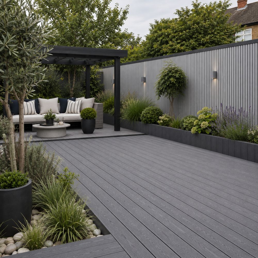 A modern outdoor patio scene with a dark gray composite decking floor, a black pergola, a gray sectional sofa, and landscaped garden beds with plants and a corrugated metal fence.